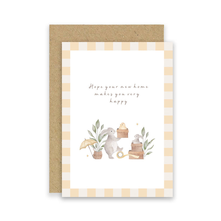 A greeting card with a kraft envelope, featuring a watercolor illustration of a bunny with a basket, small plants, and the text 'Hope your new home makes you very happy' on a white card with a beige-and-white gingham border.