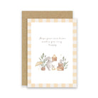 A greeting card with a kraft envelope, featuring a watercolor illustration of a bunny with a basket, small plants, and the text 'Hope your new home makes you very happy' on a white card with a beige-and-white gingham border.