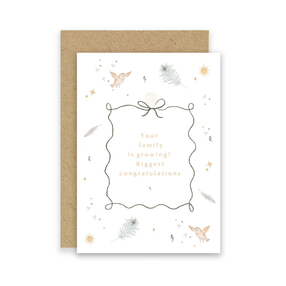 A white greeting card with a wavy border frame and gold text reading 'Your family is growing! Biggest congratulations,' shown with a brown kraft envelope behind it on a white background.